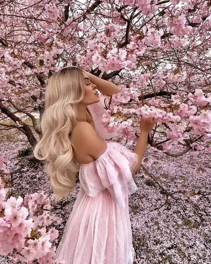 Woman in a flowing pink dress among blooming cherry blossoms, embracing nature's beauty in springtime scenery.