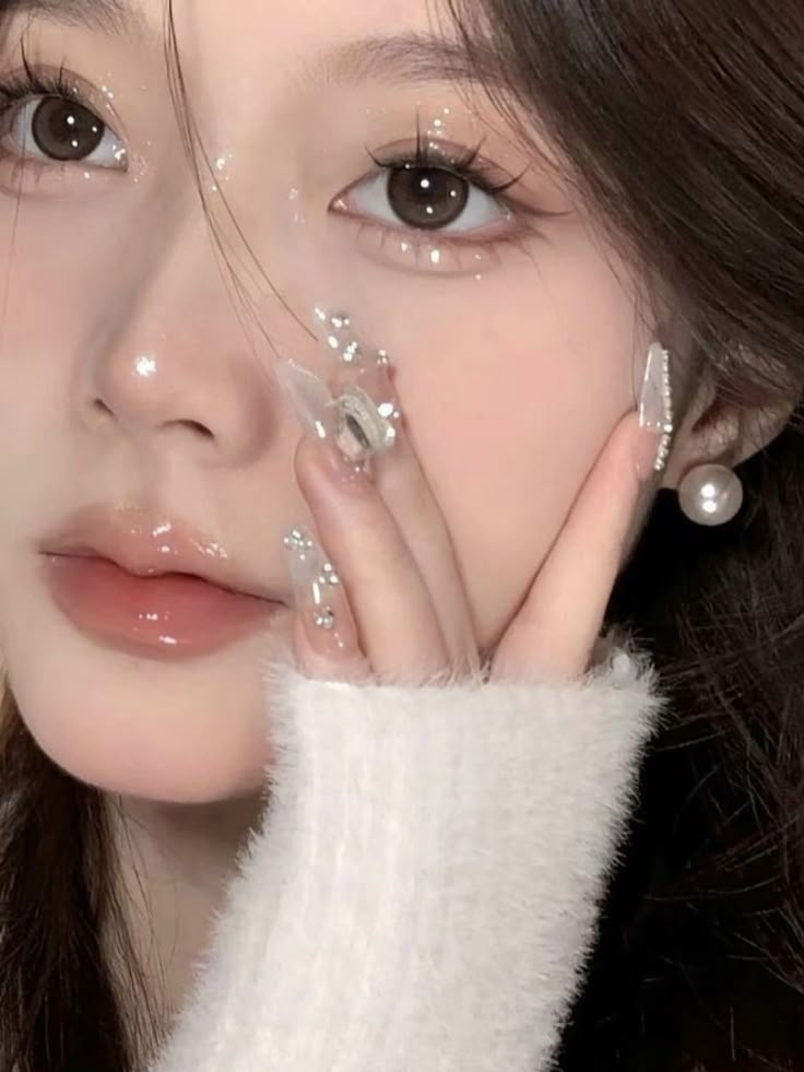 Close-up of a person with glittering makeup, glossy lips, pearl earrings, and embellished nails, wearing a soft white sweater.