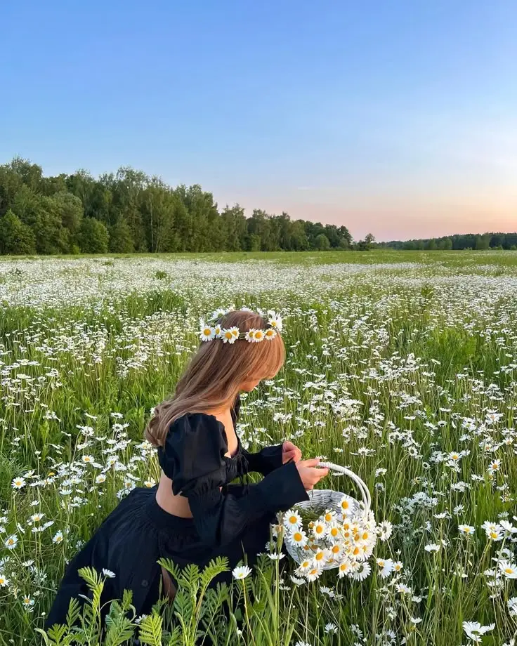 Woman with daisy crown picks flowers in a lush green field under a clear blue sky, showcasing serene nature and tranquility.