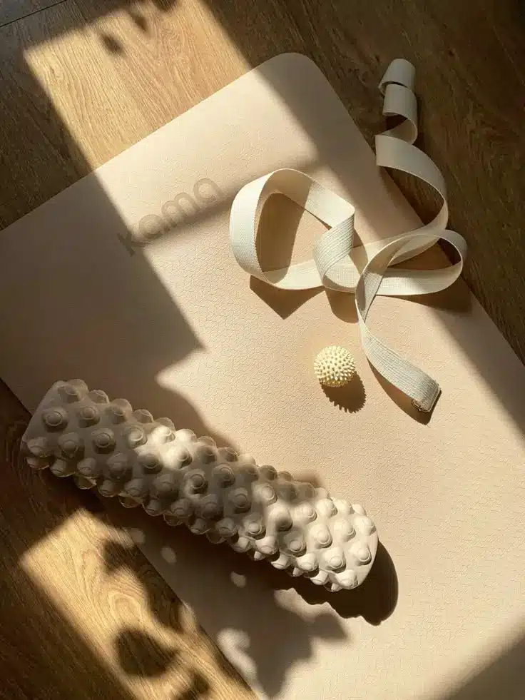 Yoga mat with a massage roller, strap, and spiky ball on wooden floor bathed in sunlight; perfect for home workout and relaxation.
