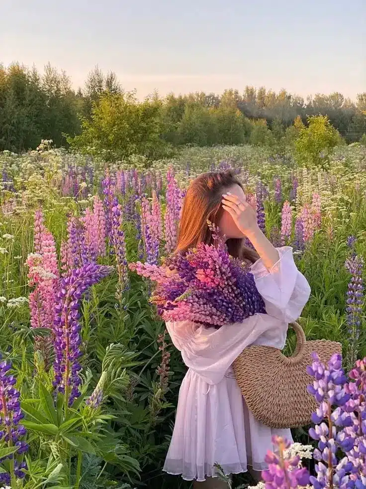 Woman in a white dress holding lupines in a sunny field, surrounded by vibrant wildflowers and lush greenery.