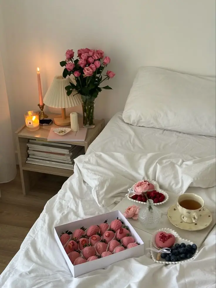 Cozy bedroom setup with pink roses, candlelit table, and a bedspread of pink-themed treats, berries, and a cup of tea.