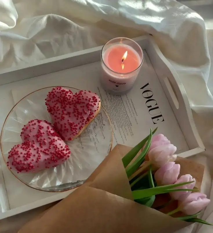Heart-shaped cookies with pink sprinkles, a lit candle, and tulips on a tray create a cozy, romantic setting.