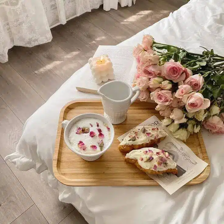 Breakfast in bed with coffee, croissants, fresh pink roses, and a candle on a wooden tray, conveying a cozy, romantic morning.