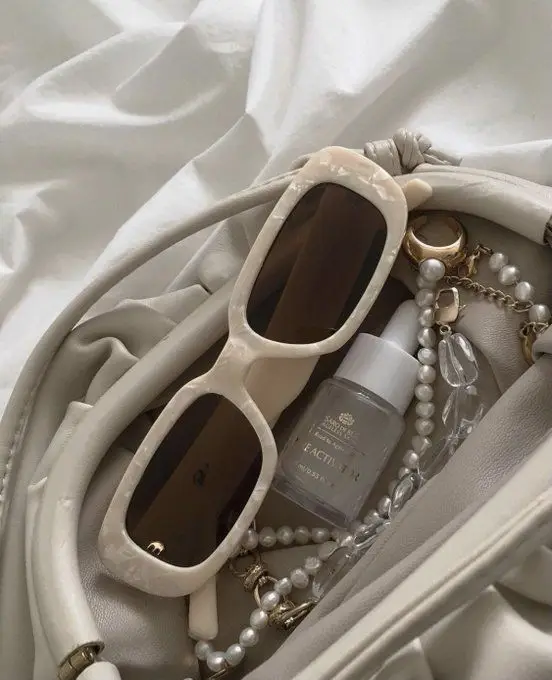 Chic beige accessories: stylish sunglasses, pearl jewelry, and luxury facial elixir on a neutral fabric background.