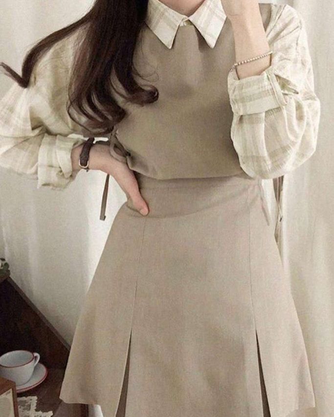 Woman in beige dress and plaid shirt, hand on hip, showcasing elegant casual fashion outfit.