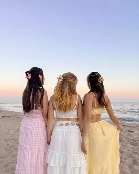 Three women in summer dresses with flower hair clips stand on a beach at sunset, facing the ocean, enjoying a serene moment.