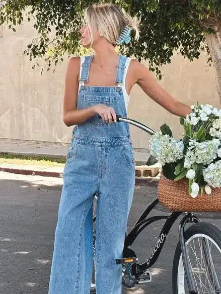 Woman in denim overalls stands beside a bike with a basket of white flowers, under a tree on a sunny day.