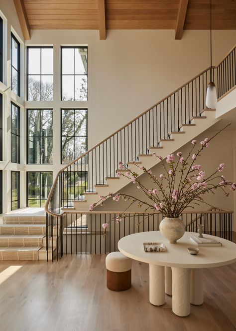 Elegant hallway with large windows, a modern staircase, and a round table adorned with a vase of blooming branches. Bright and inviting space.