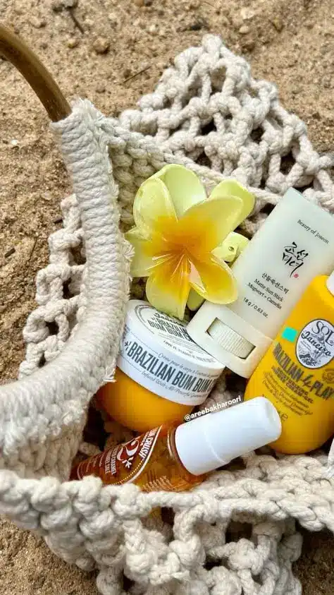 Beach essentials in a crochet bag: sunscreen, skincare products, and a yellow flower on sandy background.