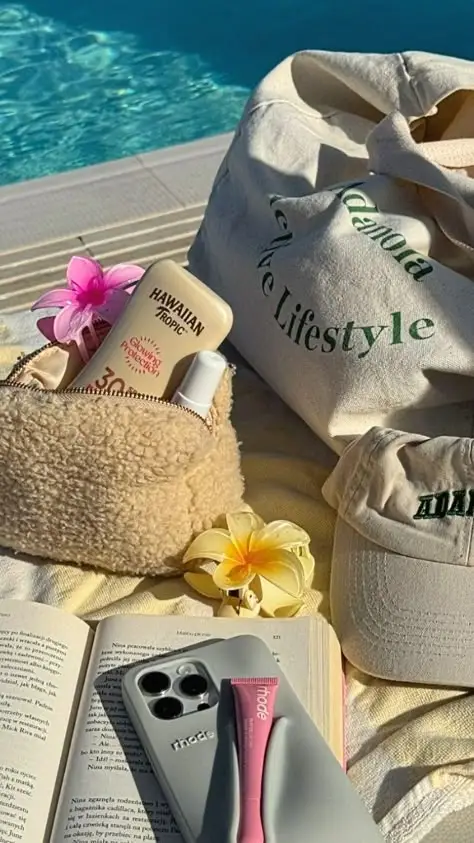 Poolside essentials: sunscreen, beige tote, casual cap, book, phone, flower, and bag with a vibrant pool backdrop.