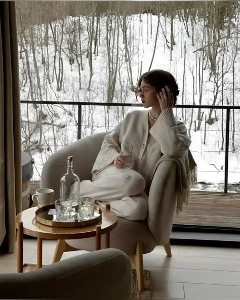 Cozy woman in a chair with a drink, gazing at snowy forest views, beside a wooden table with water bottles and glasses, indoors.