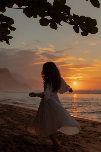 Woman in a flowing dress dances on a beach at sunset with mountains in the background, embodying freedom and tranquility.