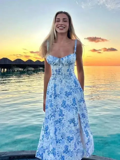 Smiling woman in a floral dress enjoying a stunning sunset by the beach, with overwater bungalows in the background.