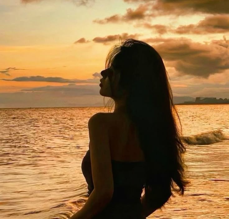 Silhouette of a woman gazing at a serene ocean sunset, with vibrant sky and gentle waves creating a peaceful beach scene.