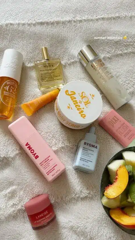 Summer beauty essentials flatlay with skincare products, body scrub, and fresh fruit on a soft white towel.