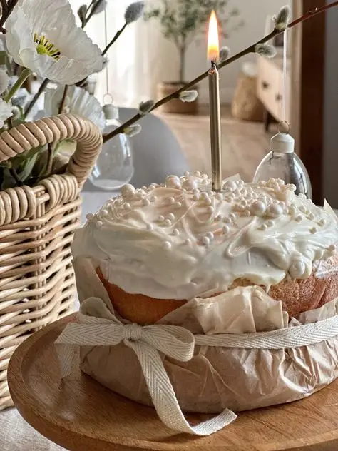 Decorative cake with white frosting and candle on wooden stand, surrounded by flowers and wicker basket, creating a cozy ambiance.
