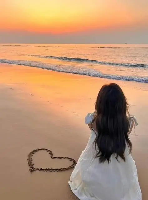 Woman in white dress sitting on beach at sunset, gazing at waves with heart drawn on sand. Serene seaside moment.