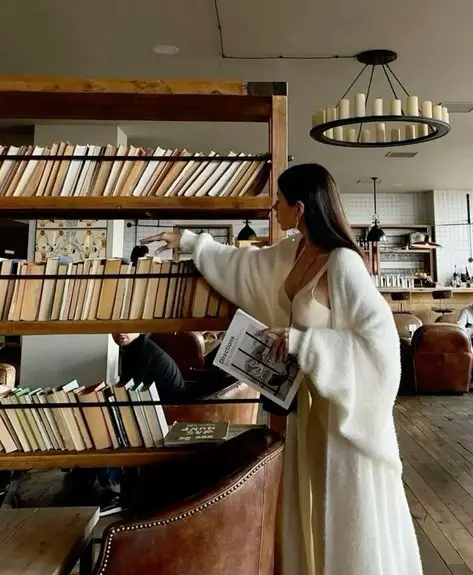 Woman in a cozy library cafe browsing bookshelves, wearing an elegant white outfit, surrounded by books and warm decor.