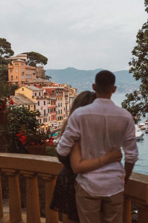 Couple embraces on a balcony overlooking a scenic coastal Italian village, with colorful buildings and boats. Romantic getaway vibes.