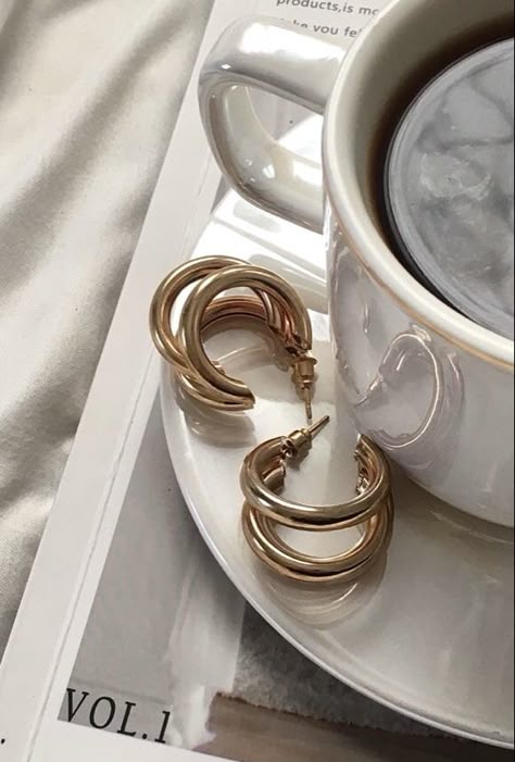 Gold hoop earrings on a magazine next to a coffee cup on a saucer, creating a stylish and cozy aesthetic.