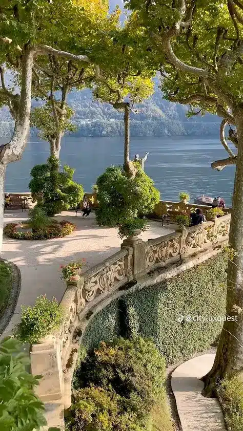 5f050795df1fd9b68db194da8d798a0a - Handy Home Scenic lakeside garden with ornate stone railing, lush greenery, and mountain views under a clear sky. Peaceful and picturesque setting.