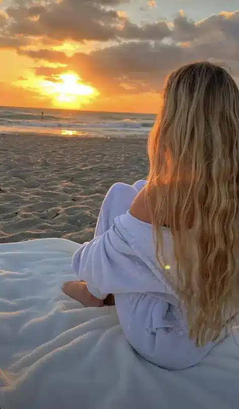 Person with long hair enjoys a serene beach sunset, sitting on a blanket. Golden light reflects on the waves and sandy shore.