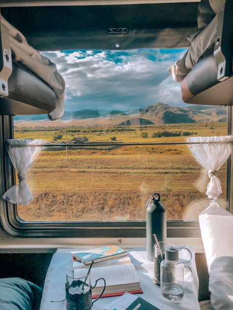 Scenic train journey view through window, showcasing mountains and field, with books and coffee on table for a cozy travel ambiance.