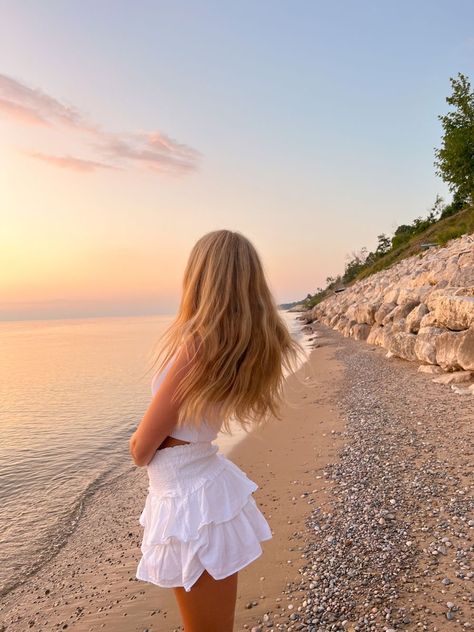 Woman in white dress gazes at serene sunset over a rocky beach, with gentle waves and a tranquil sky.