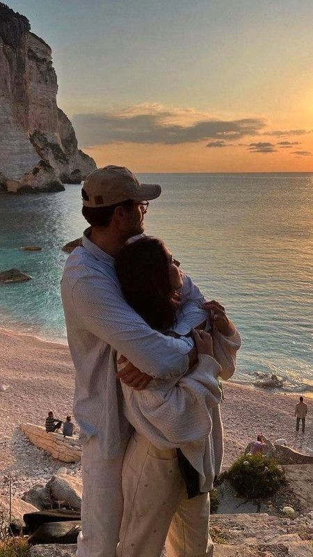 Couple embraces while watching sunset on rocky beach, serene ocean view in background. Romantic coastal scene.