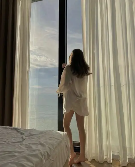 Woman in white shirt gazing out a window at serene ocean view, softly lit room, sheer curtains, peaceful morning ambiance.