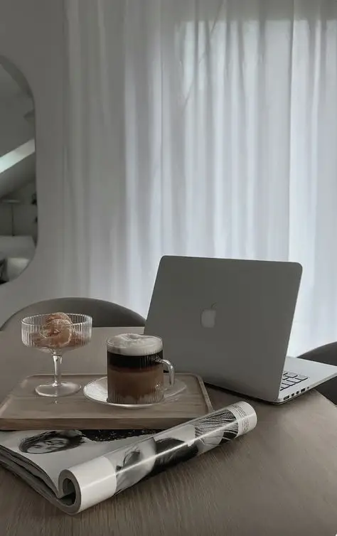 Laptop on wooden table with coffee, pastry, and magazine, set against sheer drapes. Cozy workspace vibe.