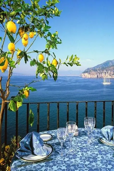 863cae4d6a9592dbfe823888e090fecc - Handy Home Table with blue cloth set for dining, overlooking ocean view with lemons hanging from a tree, ideal for a coastal dining experience.