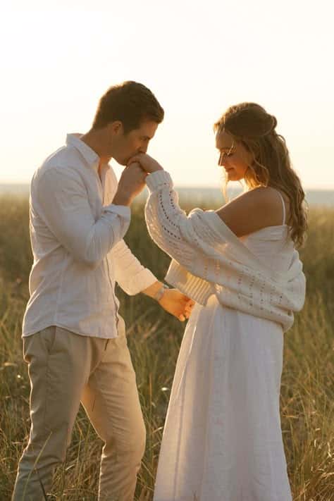 Couple in white outfits enjoying a romantic moment in a sunlit field, with gentle gestures and warm expressions.