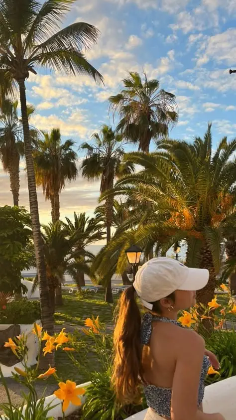 Woman in a hat enjoying a tropical garden with palm trees and flowers under a vibrant, cloudy sky at sunset.