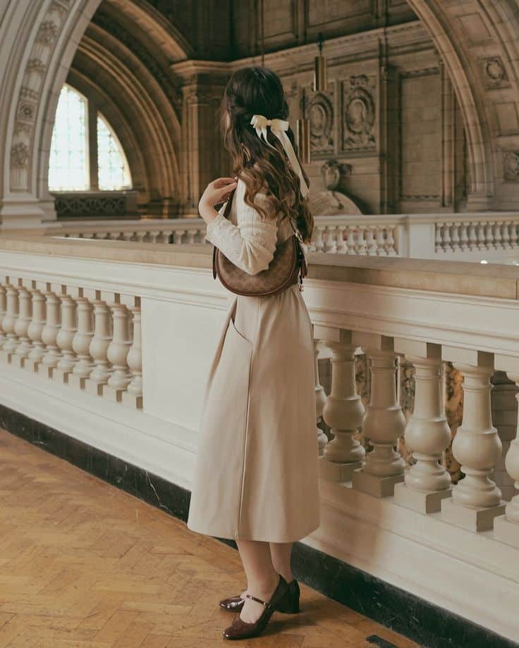 Woman in vintage outfit with ribboned hair stands on ornate balcony in historical building.