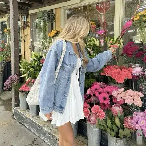 Woman in denim jacket and white dress selecting flowers at an outdoor market. Vibrant pink and green floral display.