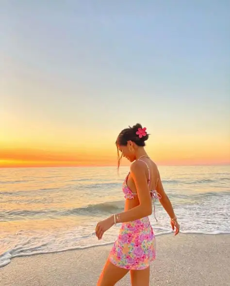 Woman in floral dress with sunset beach background, flower in her hair, standing at the ocean's edge, serene and tranquil atmosphere.