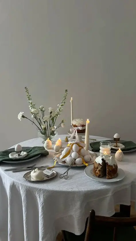 Elegant dining table setup with a white tablecloth, decorated with candles, flowers, eggs, and a dessert, creating a cozy ambiance.