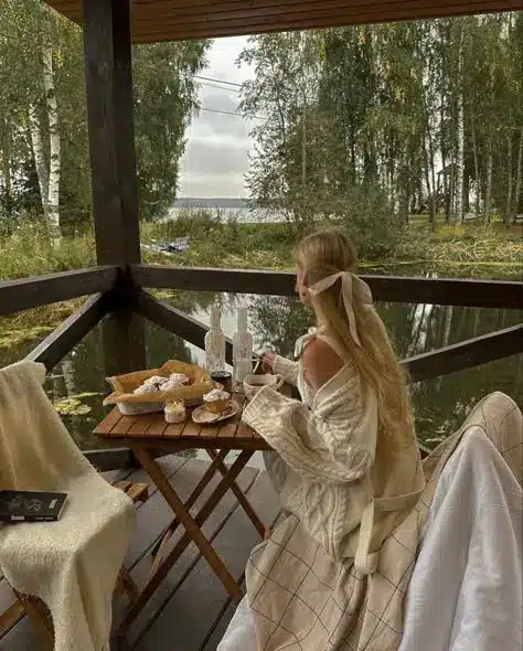 A woman enjoys coffee and pastries on a cozy porch overlooking a serene forest and lake view, wrapped in a warm blanket.