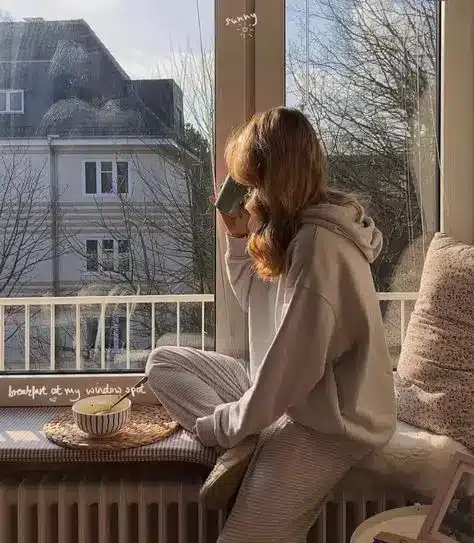 Cozy morning scene: Woman sipping coffee by a sunlit window, surrounded by pillows, with a view of a house and trees outside.