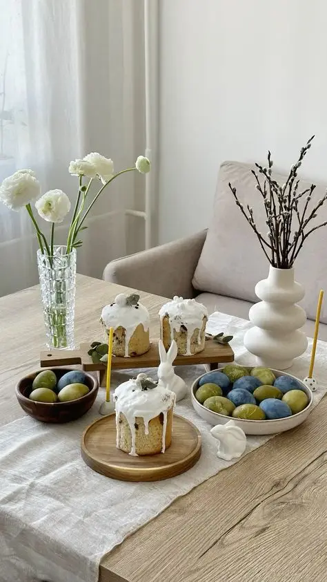 Easter table setting with decorated cakes, colored eggs, and spring flowers on a wooden table, creating a festive atmosphere.