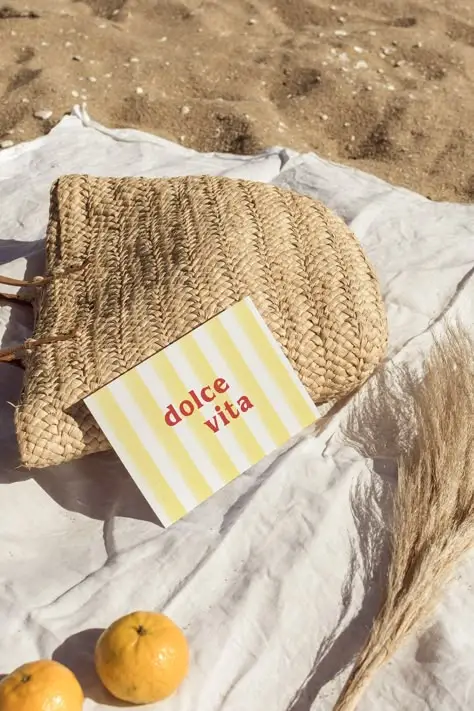 Woven beach bag on sand with dolce vita card, oranges, and pampas grass, capturing a serene summer vibe.