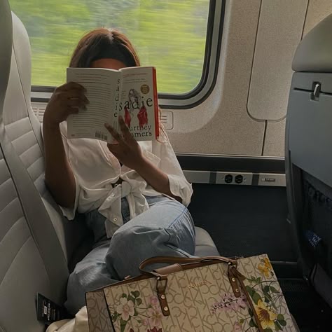 Person reading Sadie by Courtney Summers on a train, wearing casual clothes, with a floral-patterned bag nearby. Relaxed travel vibe.