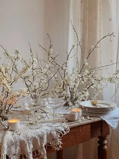 Elegant spring tablescape with white blossoms, lace tablecloth, candles, and stacked plates, creating a delicate, rustic ambiance.