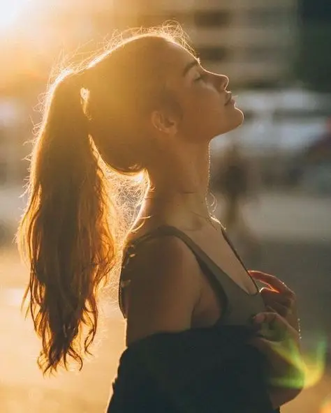 Young woman in profile soaking in golden sunset glow, hair in ponytail, peaceful expression, outdoors.