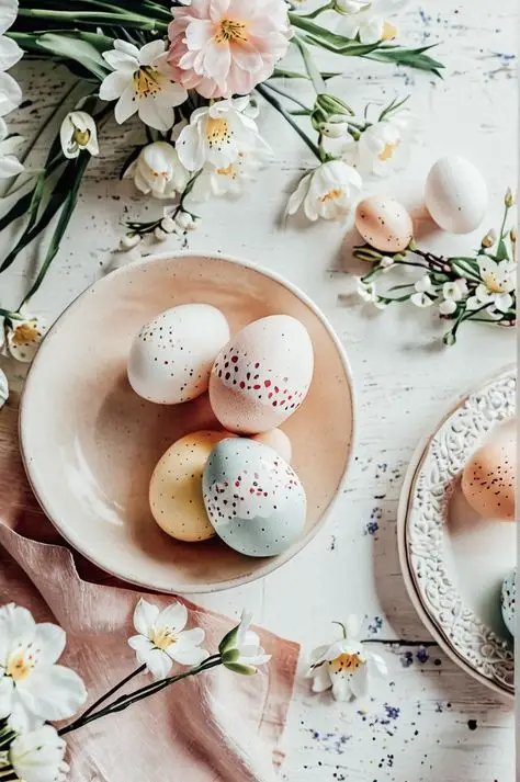 Delicate pastel Easter eggs in a bowl surrounded by spring flowers on a rustic white table.