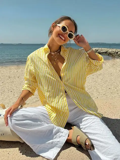 Woman in striped yellow shirt and white pants smiles on a sunny beach, wearing sunglasses. Casual summer vacation style.