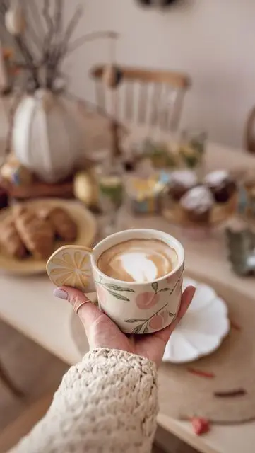 Cozy morning scene with a decorative cup of coffee in hand, pastries, and a fall-themed breakfast table setting.