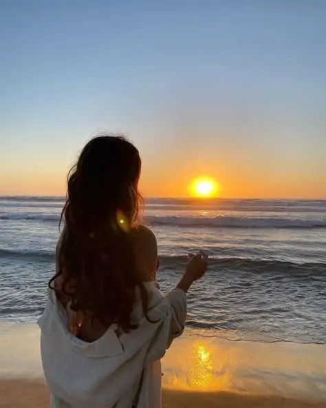 Woman enjoying a serene ocean sunset on a sandy beach, wrapped in a light sweater, with gentle waves in the background. Peaceful moment.